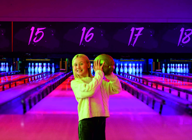 a girl holding a bowling ball in the air, in front of bowling lanes